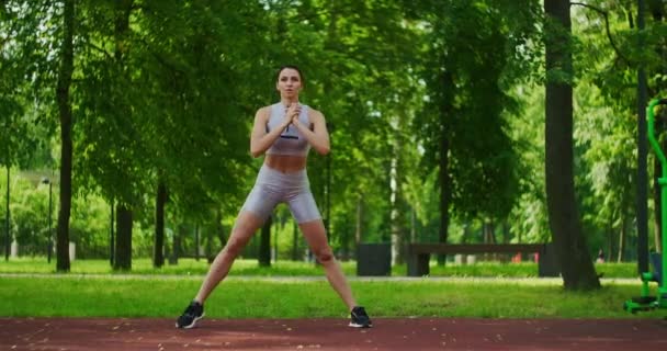 Atleta feminina executa lunges laterais exercitar suas pernas no Parque em câmera lenta. Mulher bonita jogando esportes no parque . — Vídeo de Stock