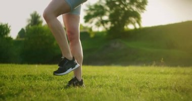 Bir kadın, geriye doğru hamle ve yüksek kalça kaldırma ile tek bacağının üzerinde çömelir. Gün batımında bacakların ve kalçaların için yaz aylarında parkta antrenman.