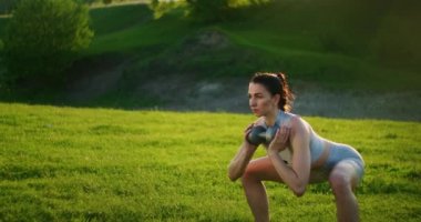 Gün batımında dambıllarla parkta çömelmiş bir kadın yan tarafa doğru hamle yapıyor. Motivasyon ve spor eğitimi. Bekar bir kadın yazın gün batımında egzersiz yapar.