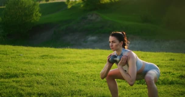 Una mujer se pone en cuclillas con pesas en el parque al atardecer realizando embestidas a un lado. Motivación y entrenamiento deportivo. Una mujer soltera hace ejercicios al atardecer en verano — Vídeos de Stock