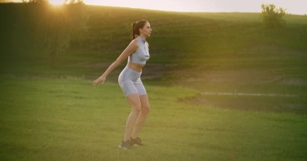 Una joven activa en un parque al atardecer se para en una pierna y salta. Se abalanza en cada pierna y salta en el Parque en el verano — Vídeos de Stock