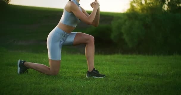 Die Kamera begleitet eine Athletin, die bei Sonnenuntergang paarweise hinunterstürzt. Bewegen Sie sich, indem Sie abwechselnd auf jedem Bein hocken. Nahaufnahme des Trainings im Park im Sommer — Stockvideo