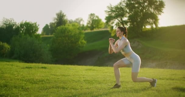 女性は後ろ向きの肺と高いヒップリフトで片方の脚でスクワットを行います。夕暮れ時の足と太もものための夏の公園でのトレーニング — ストック動画