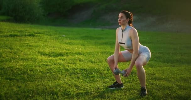 Una mujer deportiva en un parque se agacha con pesas en las manos al atardecer. Entrenamiento de pesas al aire libre. Fitness en un parque público en verano para muslos y piernas . — Vídeos de Stock