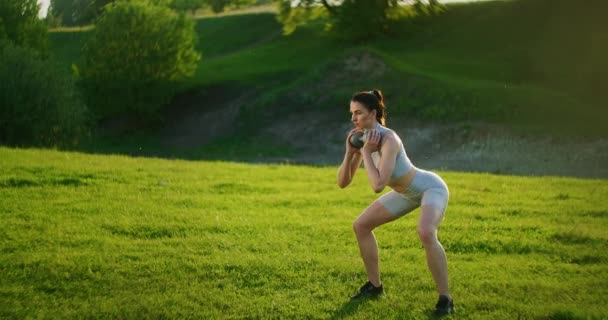 Una mujer se pone en cuclillas con pesas en el parque al atardecer realizando embestidas a un lado. Motivación y entrenamiento deportivo. Una mujer soltera hace ejercicios al atardecer en verano — Vídeos de Stock