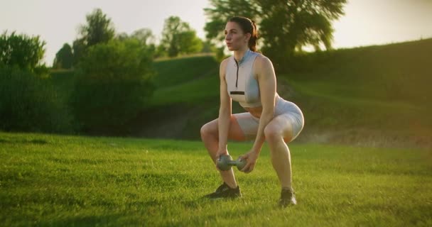 Een sportieve vrouw in een park hurkt met halters in haar handen bij zonsondergang. Gewichtstraining in de buitenlucht. Fitness in een openbaar park in de zomer voor dijen en benen. — Stockvideo