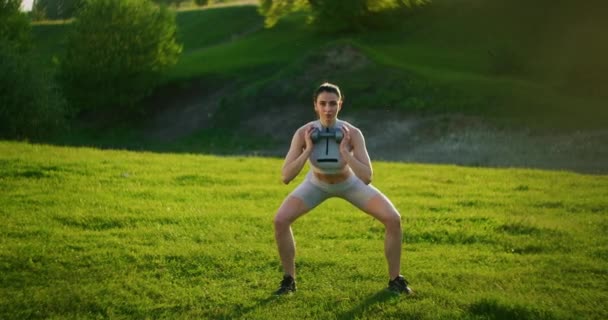 Een vrouw hurkt met halters in het park bij zonsondergang... en doet daar lunges aan de zijkant. Motivatie en sporttraining. Een alleenstaande vrouw doet oefeningen bij zonsondergang in de zomer — Stockvideo