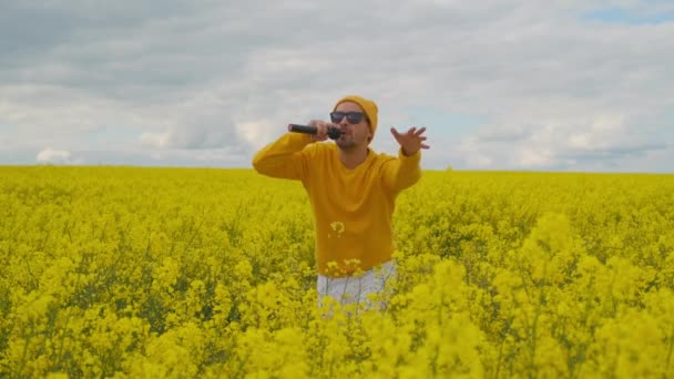 Usando óculos de sol e um campo de flores amarelas, o solista masculino em roupas amarelas dança e canta uma canção enérgica enquanto dança e acena seus braços. Câmera ao vivo — Vídeo de Stock