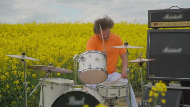 Perkusista gra na perkusji siedząc na polu rzepaku. Perkusista w kolorze żółtym — Wideo stockowe