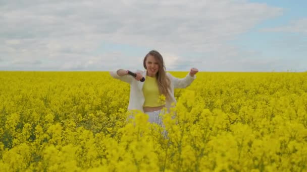 Uma cantora enérgica com um microfone em um campo de colza com flores amarelas pula balança a cabeça e dança — Vídeo de Stock