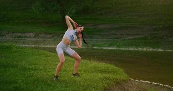 Una joven que está cerca de un río o lago. Una mujer joven mirando en las cámaras realiza inclinaciones a los lados para el entrenamiento muscular abdominal . — Vídeos de Stock