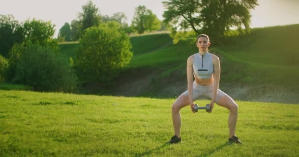 Entrenamiento de mujeres en el parque. Chica sentadillas con mancuernas en sus manos al amanecer de pie sobre la hierba — Vídeos de Stock