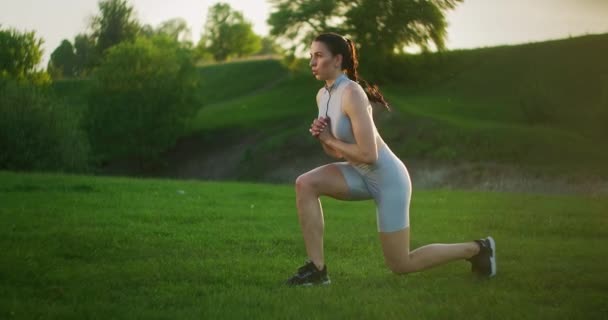 Um close-up de uma jovem mulher realiza lunges alternados em cada perna no salto. Saltar no parque ao pôr-do-sol — Vídeo de Stock