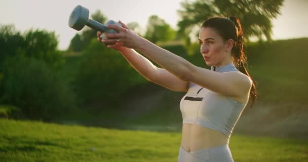 Une femme soulève un haltère dans le parc au coucher du soleil. Formation du dos — Video