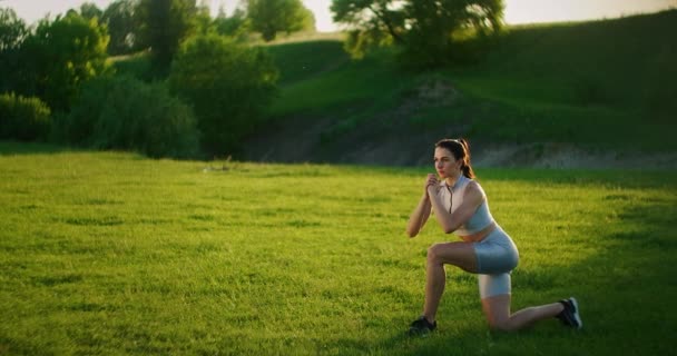 De manhã, no parque, uma mulher de sportswear executa cross-lunges para trás. Exercícios para um corpo bonito . — Vídeo de Stock