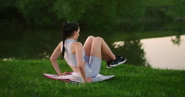 De manhã, no parque, uma mulher de roupas esportivas deitada em um tapete levanta as pernas para o corpo. Exercícios para um corpo bonito. Os músculos abdominais — Vídeo de Stock