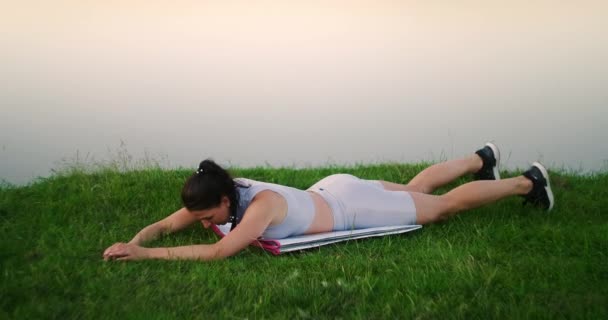 Mujer estira la espalda y hace curvas tumbadas en la hierba, chica entrena en el parque de la ciudad en la naturaleza . — Vídeo de stock