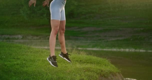 Guggol és felugrik. Egy nő gyakorolja a combja és a lába izmait a füvön a tó melletti parkban. Karcsú, gyönyörű alak. Fitness maraton. Dolgozni a testeden — Stock videók