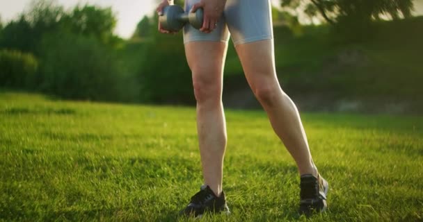 Una joven en ropa deportiva se inclina hacia adelante con pesas en la naturaleza en un parque al atardecer. Entrenamiento. Trabajar en un hermoso cuerpo por la mañana o al atardecer — Vídeo de stock
