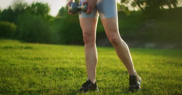 Uma jovem mulher de sportswear inclina-se para a frente com halteres na natureza em um parque ao pôr do sol. Treino. Trabalhe em um belo corpo de manhã ou ao pôr do sol — Vídeo de Stock