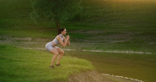 Kraakt en springt omhoog. Een vrouw doet oefeningen voor de spieren van haar dijen en benen op het gras in een park in de buurt van het meer. Slank, mooi figuur. Fitnessmarathon. Werken aan je lichaam — Stockvideo