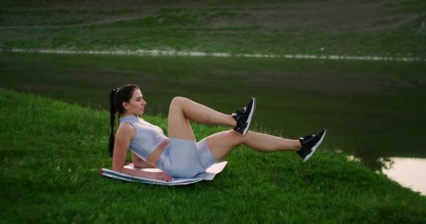 Uma mulher faz exercícios para os músculos da imprensa deitada na grama em um parque perto do lago. Trabalhar com músculos abdominais. Criando um corpo bonito — Vídeo de Stock