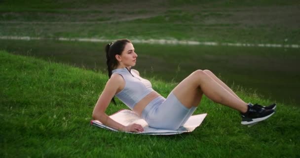 Eine Frau macht in einem Park in der Nähe des Sees Übungen für die Muskeln der Presse, die auf dem Rasen liegen. Arbeiten Sie mit Bauchmuskeln. Schaffung eines schönen Körpers — Stockvideo