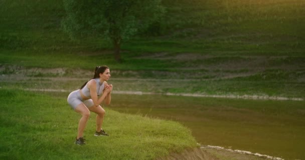 Lunges in the side. A woman does exercises for the muscles of her thighs and legs on the grass in a Park near the lake. Work with abs muscles. Slim, beautiful figure. Fitness marathon — Stock Video