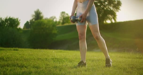 Молодая женщина в спортивной одежде наклоняется вперед с гантелями на природе в парке на закате. Тренировка. Работа на красивом теле утром или на закате — стоковое видео