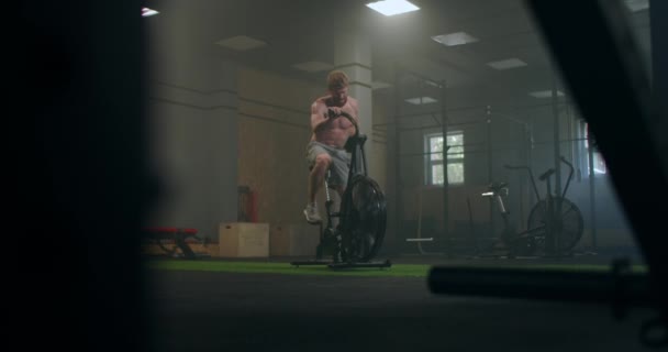 Un homme robuste surmontant les trains de fatigue sur un vélo stationnaire dans une salle de fitness. gymnase atmosphérique. athlète de secours — Video