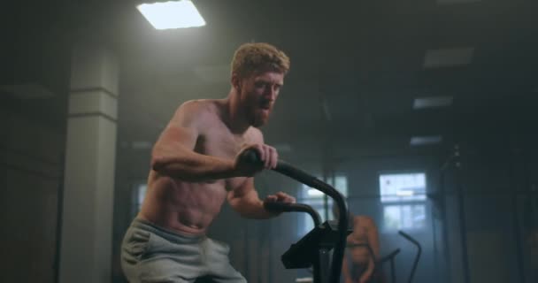 Un hombre resistente que supera los trenes de fatiga en una bicicleta estacionaria en una sala de fitness. gimnasio atmosférico. atleta de alivio — Vídeos de Stock