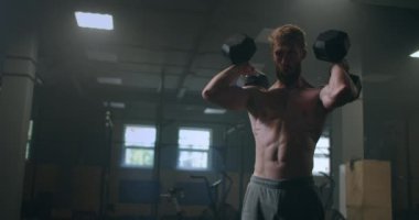 Tişörtsüz bir adam spor salonunda dambıl kaldırıyor. Cesur bir atlet.