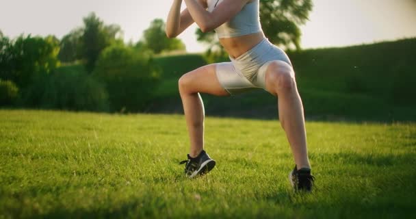 Uma jovem realiza um exercício estático na grama para os músculos da perna e quadril. Levantar-se em dedos dos pés enquanto sentado. Exercício para a perna inferior. Treinamento na rua no Parque — Vídeo de Stock