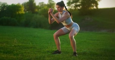 Genç bir kadın gün batımında çimlerin üzerindeki parkta çadır egzersizinden atlıyor. Çayırda genç bir kadının yavaş çekim fitness eğitimi