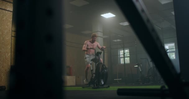 Молодий чоловік використовує тренувальний велосипед у спортзалі. Фітнес-чоловік використовує повітряний велосипед для кардіо-тренувань у спортзалі. Обладнання, зусилля . — стокове відео