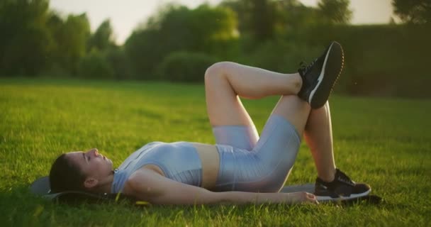 Una giovane donna sdraiata nel parco sull'erba su un tappetino sportivo esegue ascensori pelvici. Esercizi per i muscoli delle natiche. Esercizi con il proprio peso senza pesi. — Video Stock