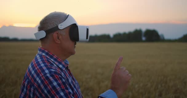 Älterer erwachsener Bauer in einem Virtual-Reality-Helm auf einem Getreidefeld. Im Sonnenuntergang bedient sich ein älterer Mann in einem Traktorfahrer einer Virtual-Reality-Brille. VR-Technologien und modernes Agrargeschäft — Stockvideo