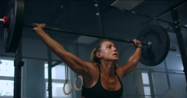 Langzame beweging: Training squats met halters over het hoofd. Gespierde vrouwelijke heffen lange halter over hoofd training schouders in de sportschool. Zelfverzekerde fitnessvrouw die krachttraining beoefent met zwaar materieel. — Stockvideo