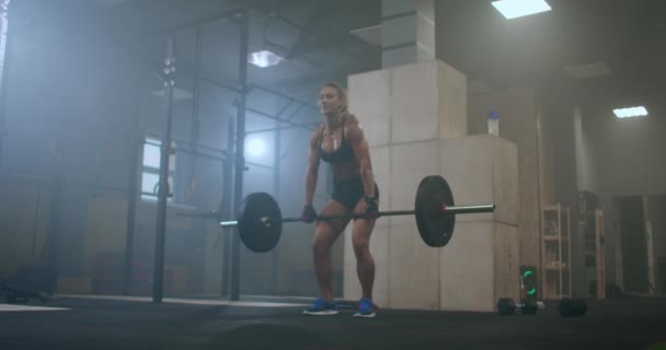 Una mujer levantadora de pesas realiza un levantamiento de pesas en un gimnasio oscuro. una mujer levantando una barra pesada sobre su cabeza — Vídeos de Stock