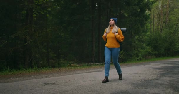 Vista frontale, pianta principale: una donna che viaggia a piedi in maglione giallo e cappello su una strada forestale con uno zaino e vista sugli alberi e la natura. Da solo in viaggio. Il concetto di libertà e di viaggio. — Video Stock