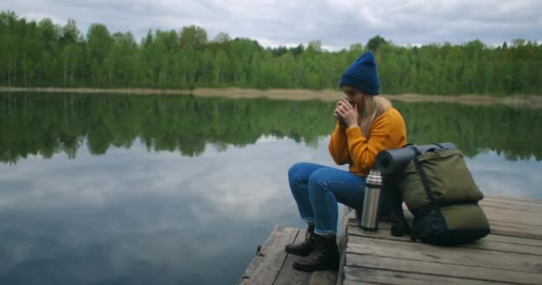 帽子とリュックを被った女性が山の中の森の湖の木製の桟橋に座って、魔法瓶から熱い飲み物茶やコーヒーを飲みます. — ストック動画