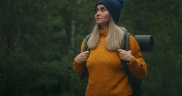 Retrato baixo ângulo uma mulher com uma mochila em um suéter amarelo e chapéu está andando ao longo de uma estrada na floresta em câmera lenta . — Vídeo de Stock
