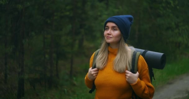 Una donna viaggia a piedi in maglione e cappello lungo una strada forestale con uno zaino ed esamina gli alberi e la natura. Da solo in viaggio. Il concetto di libertà e di viaggio. — Video Stock