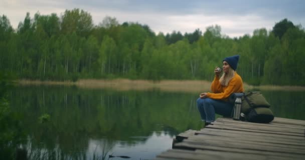 女性の旅行者はバックパック付きのお茶を飲み、木製の桟橋に座っている山の森の湖の美しさを楽しんでいます。孤独な女性の孤独な旅のスローモーションの概念。隔離と旅行 — ストック動画