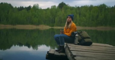 Sırt çantalı genç bir kadın baharda gün batımında dağ gölünün kıyısında oturuyor. İtalya 'ya git. Zayıf kızla manzara, sudaki yansıma, yeşil ağaçlar. Sırt çantalı Bayan Turist