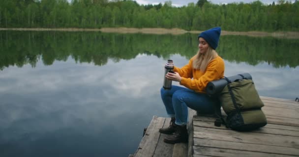 Uma viajante do sexo feminino bebe chá com uma mochila e desfruta da beleza de um lago de floresta montanhosa sentado em um cais de madeira. Conceito de câmera lenta de uma viagem solitária de mulheres solitárias. Quarentena e viagens — Vídeo de Stock