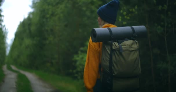 Vue arrière du bas et du côté : une femme voyageant à pied dans un pull jaune et un chapeau sur une route forestière avec un sac à dos et regardant les arbres et la nature. Seul en voyage. Le concept de liberté — Video