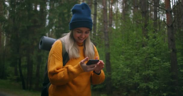 黄色のセーターとバックパック付きの帽子の女性が携帯電話を持っていて、メッセージを入力しています。画面上で指をタップし、笑顔。道路上の森の中の旅行者はスマートフォンを使用しています — ストック動画