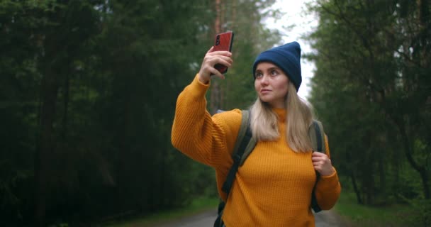 O femeie își face un selfie pe telefon și călătorește pe jos într-un pulover galben și o pălărie de-a lungul unui drum forestier cu un rucsac. Raport foto și blog de călătorie — Videoclip de stoc