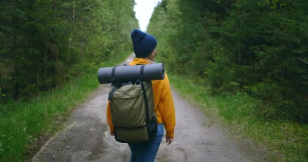 Вид ззаду жінка з рюкзаком в жовтому светрі і капелюсі, що йде по дорозі в лісі в повільному русі . — стокове відео